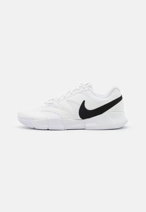 Bílé atletické boty se strukturovaným svrškem, černým logem Nike, polstrovaným límcem a pevným gumovým podešvem s vzorovaným profilem.