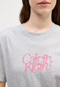Harmaa puuvillapaita, jossa on suuri vaaleanpunainen logo, jossa lukee "Calvin Klein" tyylitellyssä fontissa. Paidassa on pyöreä kaula-aukko ja lyhyet hihat.