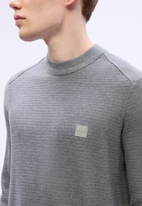 Pulover gri tricotat cu un finisaj texturat, având un gât rotund și un mic patch logo pătrat pe piept.