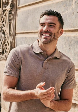 Lachende man in een bruin poloshirt staand buiten tegen een stenen muur, handen gevouwen en naar de zijkant kijkend.