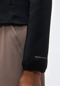 Κοντινό πλάνο του χεριού ενός ατόμου που φοράει μαύρο μπουφάν με την επιγραφή "OMNI-WICK" στο μανίκι και μπεζ παντελόνια με ορατή ραφή και τσέπη.