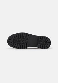 Semelle de botte noire avec des crampons profonds présentant des motifs croisés et rectangulaires pour une bonne traction sur un fond blanc uni.