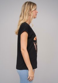 Svart bomulls-t-skjorte med rullede ermer, med blomster grafisk trykk foran. Modellen har på seg blå denim jeans. Enkelt design, myk tekstur.