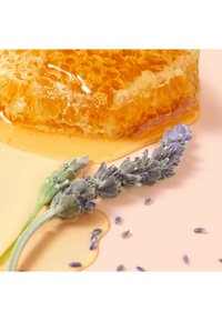 Kawałek plastra miodu o złoto-żółtym kolorze i teksturowanej powierzchni, w towarzystwie świeżych gałązek lawendy, na jasnoróżowym tle.