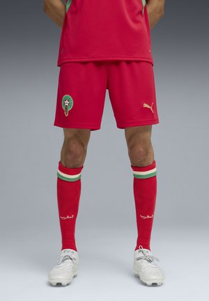 Jugador de fútbol masculino que lleva pantalones cortos rojos con el emblema de Marruecos, calcetines rojos a juego con rayas verdes y blancas, y botas blancas.