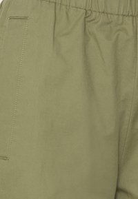 Zbliżenie na spodnie z elastycznym pasem w oliwkowym kolorze, z widocznym szwem bocznym i przeszytym otwarciem kieszeni.