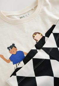 T-shirten i crème bomull har lekfulla tryckta figurer. Svart och vit rutigt tyg är lagrat under, vilket skapar kontrast i designen.