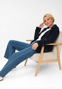 Marine uldblandingsjakke, hvid bluse og blå denimjeans. Sidder på en stribet beige stol med træsben, hånd på hagen.