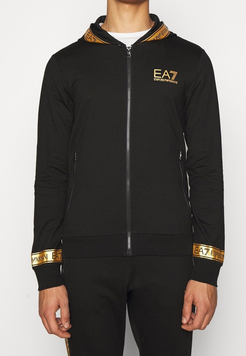 Schwarze Zip-Hoodie mit einem kontrastierenden goldenen Logo und bedruckten Akzenten an Kapuze und Bündchen, aus glattem, dehnfähigem Material.