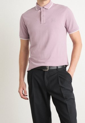 Polo violet clair avec col classique, deux boutons et bordure blanche. Porté avec un pantalon noir plissé et une ceinture noire.