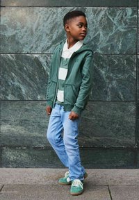 Jongen in een groene hoodie, gestreepte polo, lichtblauwe spijkerbroek en groene sneakers staat naar de zijkant te kijken tegen een donkere stenen muur.
