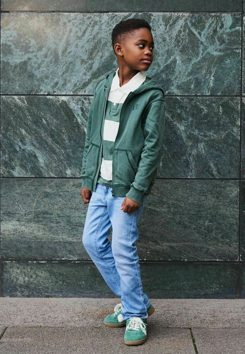Jongen in een groene hoodie, gestreepte polo, lichtblauwe spijkerbroek en groene sneakers staat naar de zijkant te kijken tegen een donkere stenen muur.