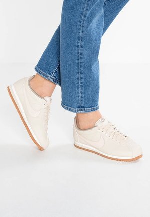 Person iført beige Nike-sneakers og blå jeans, der står på en hvid baggrund med den ene fod let hævet.