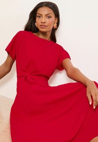 Červené šaty s krátkymi rukávmi, detailom viazania v páse a voľnou sukňou. Hladká látka v jednofarebnom prevedení, minimalistický dizajn.