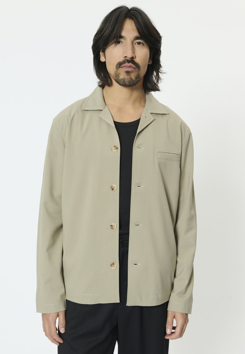 Bézs színű, hosszú ujjú ing, galléros nyakkivágással, elöl gombos lezárással és egyetlen mellzsebbel; sima anyagból készült. Látható fekete aláöltözet.