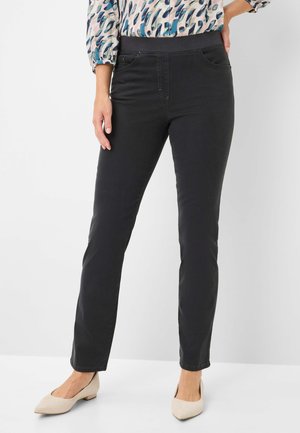 Frau trägt schwarze gerade geschnittene Hose mit elastischem Bund, gemustertes langärmeliges Oberteil und beigefarbene, flache Schuhe, steht mit der Hand in der Hüfte.