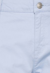 Vaaleansininen denimmateriaali, jossa on suora leikkaus, kontrastina valkoiset tikkausaumat ja hopeinen metallinen nappikoriste vyötäröllä.