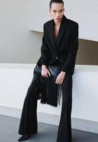 Blazer sartoriale nero con profonda scollatura a V, abbinato a pantaloni a gamba larga. Tienete una borsa in pelle nera con dettagli a frange. Tessuto liscio.