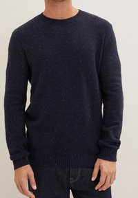 Man som bär en mörk marinblå spräcklig stickad tröja med rund halsringning, långa ärmar och ribbad avslutning vid ärmarna och nederkanten.