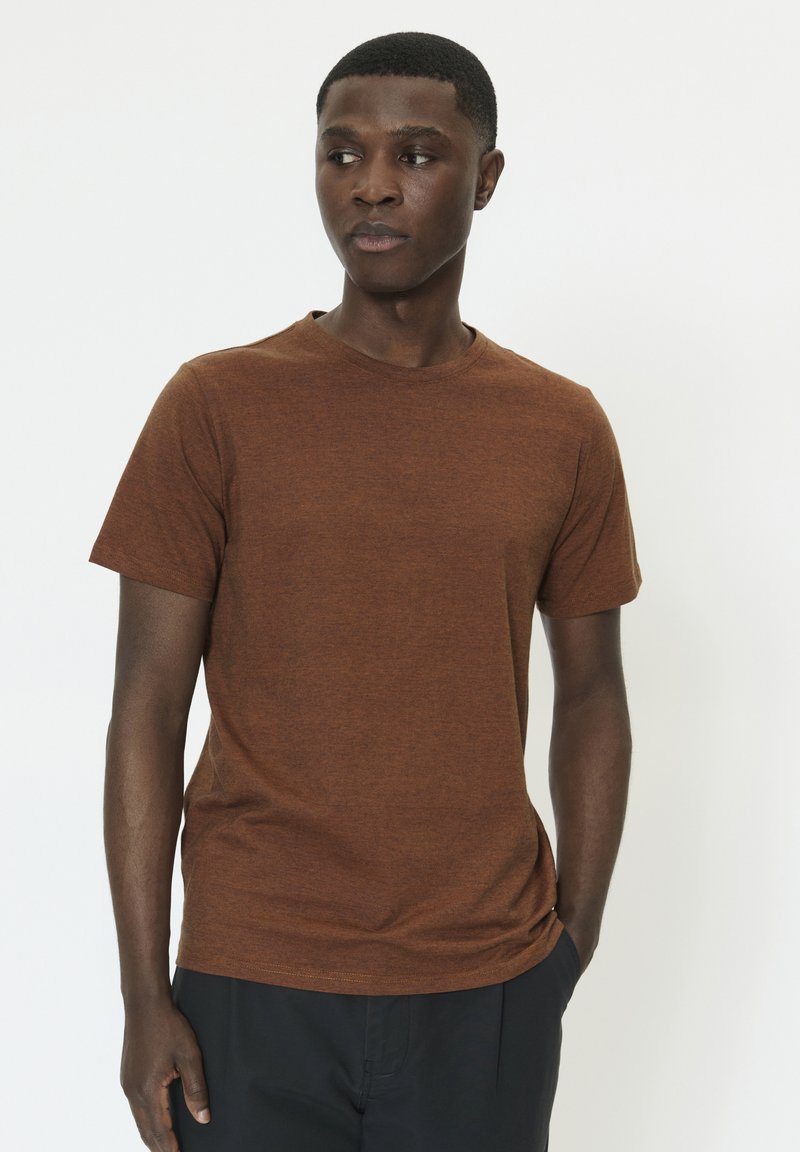 T-shirt marron en bruyère à manches courtes avec un col rond et une finition douce et texturée. Porté avec un pantalon noir, sur un fond blanc.
