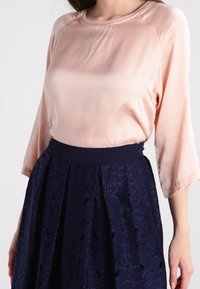 Vaaleanpunainen satiinik blouse, jossa on pyöreä pääntie ja kolmannesosa hiharat, yhdistettynä tummansiniseen pitsihameeseen, jossa on levenevä muoto ja kukkakuvioita.