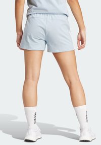 Person in hellblauen Sportshorts, weißen Adidas-Socken und weißen Sneakers, steht mit leicht gespreizten Beinen und zeigt mit dem Rücken.