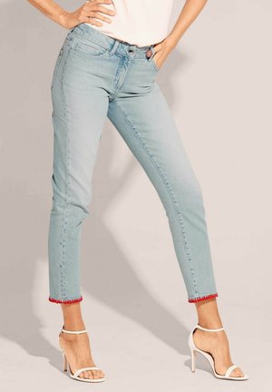 Lichtblauwe denimjeans met een cropped snit, voorzien van een rode pomponrand aan de manchetten. Gecombineerd met witte hakken met bandjes.