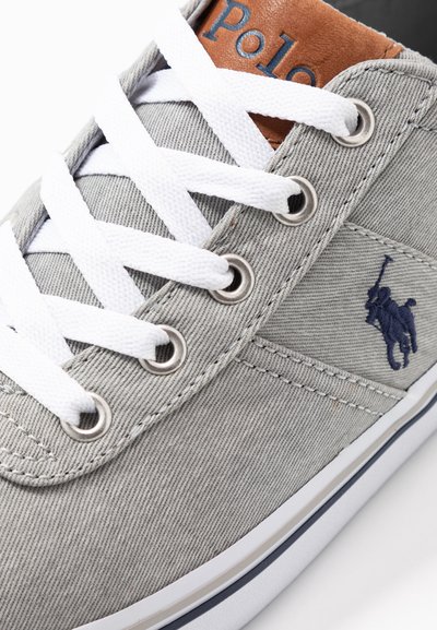 Sapatilha de lona cinza com atacadores brancos, ilhós prateados e um logo azul da polo. Acentuada com um colarinho de couro castanho e sola branca.