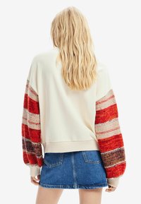 Kremowy sweter z wielokolorowymi, dzianinowymi rękawami z czerwonymi i brązowymi paskami, w połączeniu z dżinsową mini spódnicą.