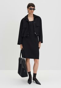 Donna che indossa una giacca nera a maniche corte, un vestito nero senza maniche, calze nere trasparenti, scarpe nere a punta, occhiali da sole, mentre tiene una borsa in rete nera.