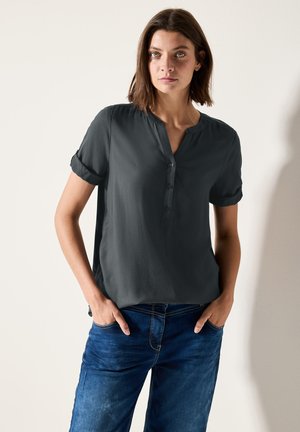 Dunkelgraues kurzärmliges Blusenshirt mit einem Henley-Kragen und drei Knöpfen, mit umgeschlagenen Ärmeln und lockerem Schnitt, kombiniert mit blauen Jeans.