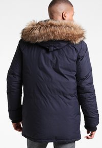 Marineblå pufferjacka med foder av konstpäls vid huvan, quiltad struktur och elastiska ärmslut. Designen är midlång med rak skärning.