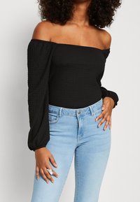 Musta olkaimeton paita, jossa on tekstuuri, pitkät pallohihat, yhdistettynä vaaleansinisiin korkeavyötäröisiin farkkuihin, joissa on hopeinen rengas.