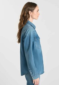 Kvinna med långt brunt hår som bär en blå denimskjorta med knappar och matchande jeans, stående i profil mot en enkel bakgrund.