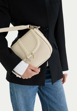 Person wearing black blazer and blue jeans holds a beige leather shoulder bag with a strap and buckle detail.