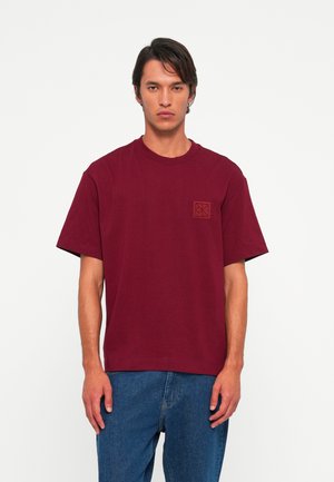 Man wearing a plain burgundy short-sleeve T-shirt with a small chest logo, paired with loose blue jeans, standing against a white background.