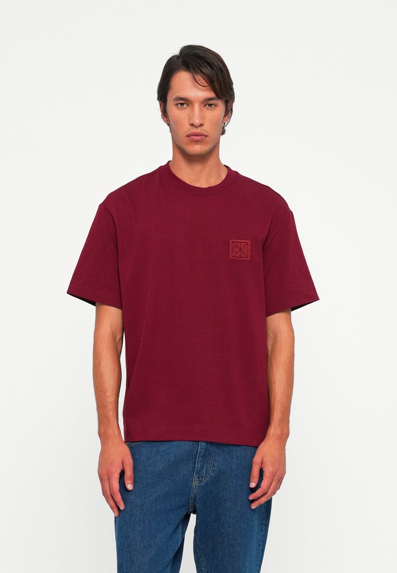 Hombre vistiendo una camiseta de manga corta burgundy lisa con un pequeño logotipo en el pecho, combinada con jeans azules holgados, de pie contra un fondo blanco.