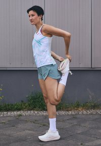 Camiseta atlética ligera con patrones azules, combinada con pantalones cortos verde claro. Zapatillas blancas y calcetas hasta el tobillo. Posición de estiramiento contra un fondo gris.