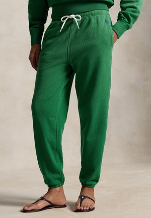 Άνδρας που φοράει πράσινο παντελόνι φόρμας με κορδόνι και μπλε σαγιονάρες, στέκεται με το ένα χέρι στην τσέπη μπροστά σε ουδέτερο φόντο.