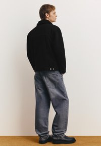 Mladi moški v črni jakni z ovčjim ovratnikom, ohlapnih sivih kavbojkah in črnih škornjih stoji bokom ob beli steni.