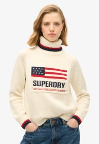 Maglione a collo alto color crema con un prominente design della bandiera americana e il testo "SUPERDRY", con accenti a coste rossi e blu navy.