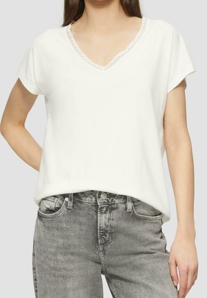 Femme portant un haut blanc à manches courtes avec décolleté en V orné de dentelle et un jean gris taille haute, debout, une main derrière le dos.