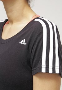 Camiseta deportiva negra con escote redondo, con tres rayas blancas en cada hombro y un pequeño logo blanco de Adidas en el pecho.
