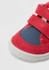 Baskets en daim rouge avec un accent en maille bleue, dotées de deux bandes Velcro et d'une semelle en caoutchouc texturée. Vue rapprochée de la zone des orteils.