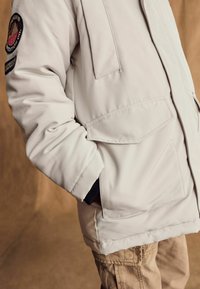 Geacă izolată bej, cu un buzunar frontal și un patch brodat pe mânecă. Mânecile cu mansetă au o margine din tricot negru.
