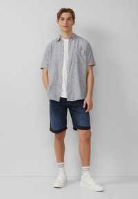 Korte mouwen, overhemd met knopen in grijs met een subtiel patroon, gecombineerd met omgeslagen marineblauwe shorts en witte sneakers met gestreepte sokken.