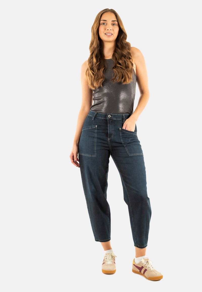 Pantalones de mezclilla azul oscuro con bolsillos, diseño de pierna ajustada; combinados con una blusa sin mangas de color gris metálico y deportivas beige con acentos rojos.
