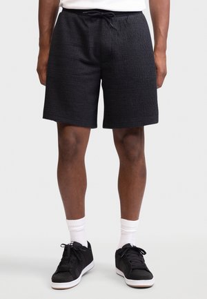 Homme portant un short noir texturé à cordon, des chaussettes blanches et des baskets noires, debout devant un fond uni.