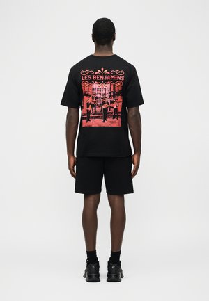 Muž oblečený v čiernych šortkách a čiernom tričku, na chrbte s červenou grafikou štyroch hudobníkov a textom "LES BENJAMINS".