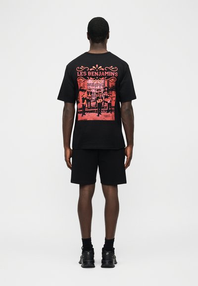 Homme portant un short noir et un T-shirt noir avec un graphisme rouge représentant quatre musiciens et le texte "LES BENJAMINS" au dos.